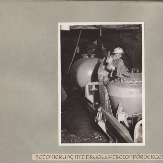 Ein historisches Bild eines Mannes, der ein großes mechanisches Gerät in einem Bergwerk bedient, mit großen zylindrischen Tanks im Hintergrund.