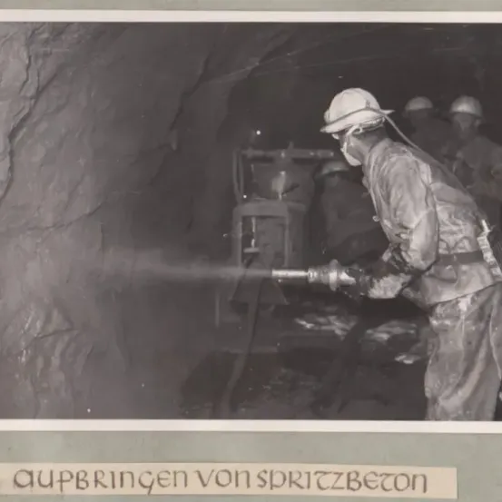 Ein Arbeiter mit Helm und Schutzkleidung spritzt Wasser in einem Tunnel. Andere Arbeiter stehen in der Nähe, und die Tunnelwände sind mit Felsen bedeckt.