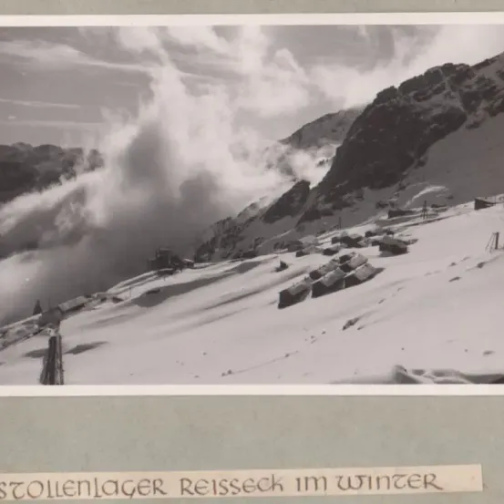 Ein schwarz-weißes Foto zeigt eine Winterlandschaft mit schneebedeckten Bergen, dichtem Nebel und einem Dorf in der Ferne. Die Szenerie ist ruhig und friedlich, mit einer Atmosphäre der Stille.