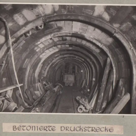 Ein Bild eines Tunnels mit Ziegelwänden und Betondecke. Ausrüstung und Werkzeuge sind verstreut an den Seiten zu sehen. Ein deutsches Wort 'BETONIERTE DRUCKSTRECKE' befindet sich am unteren Rand.
