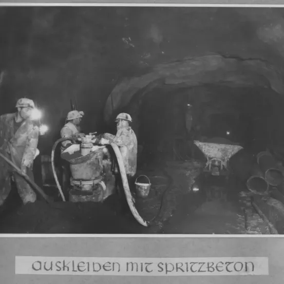 Ein Schwarz-Weiß-Foto von Arbeitern in einem Tunnel. Ein Arbeiter benutzt eine Maschine, ein anderer hält eine Schaufel, und ein dritter arbeitet mit einem Schlauch. Der Tunnel hat Rohre und Ausrüstung.