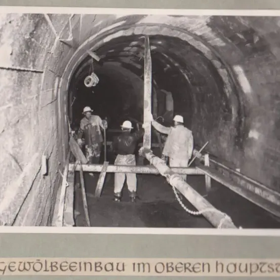 Schwarz-Weiß-Foto von Arbeitern in einem Tunnel mit einem großen Pfahl in der Mitte. Arbeiter tragen Helme und Arbeitskleidung. Der Tunnel ist eng und schwach beleuchtet.