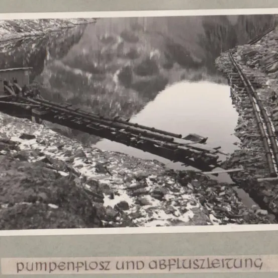 Ein Schwarz-Weiß-Foto zeigt einen Fluss mit einer beschädigten Brücke, Schutt und darunter den Text 'pumpenplossz und abflusszlerung'.