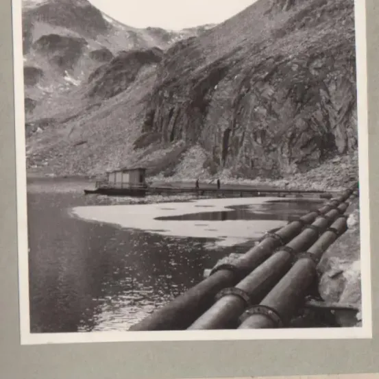 Ein Schwarz-Weiß-Foto zeigt eine Berglandschaft mit einem Gewässer. Ein kleines Boot schwimmt auf dem Wasser, und mehrere Rohrleitungen erstrecken sich vom Ufer. Zwei Personen stehen in der Nähe des Bootes. Die Bildunterschrift lautet 'schwimmende pumpanlage.'