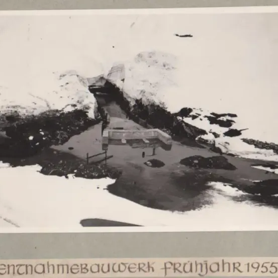 Ein Schwarz-Weiß-Foto zeigt eine Baustelle mit schneebedeckter Umgebung aus den frühen 1950er Jahren. Ein teilweise errichtetes Gebäude, umgeben von felsigem Gelände, ist sichtbar.