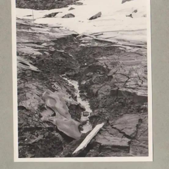 Ein Schwarz-Weiß-Foto zeigt eine raue Landschaft mit einem schmelzenden schneebedeckten Berg im Hintergrund und einem rissigen, felsigen Gelände im Vordergrund.