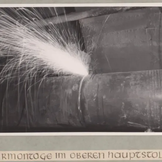 Ein Schwarz-Weiß-Foto einer industriellen Umgebung zeigt Dampf oder Gas aus einer Rohrleitung. Das Rohr ist groß und horizontal positioniert.