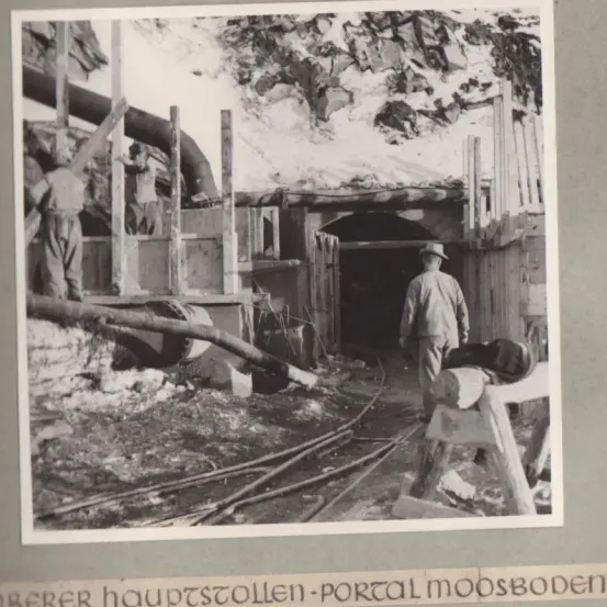 Ein Schwarz-Weiß-Foto einer Gruppe von Arbeitern in einem Tunnel, einige mit Hüten und Werkzeugen. Der Tunnel ist eng und mit Schutt gefüllt.