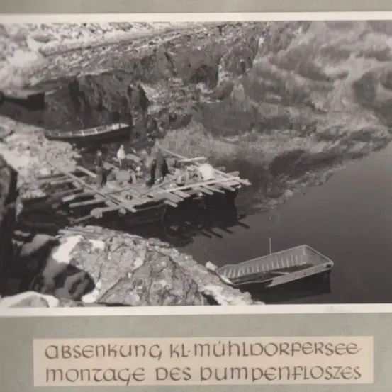 Schwarz-Weiß-Foto, das eine Gruppe von Menschen auf einer Holzplattform in einem Fluss zeigt, mit einem angedockten Boot und einer Bildunterschrift, die 'Absenkung Mühldorfersee, Montage der Pumpenschleusen' lautet.