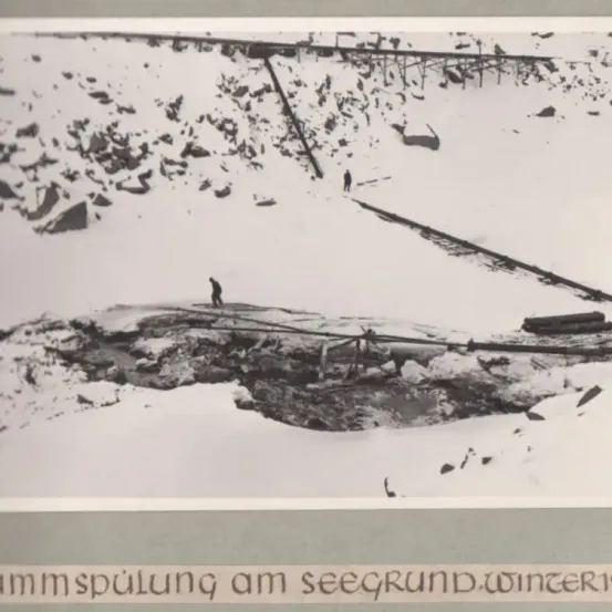 Ein Schwarz-Weiß-Bild zeigt eine verschneite Berglandschaft mit Felsen und einer beschädigten Eisenbahnstrecke. Zwei Personen gehen im Schnee.