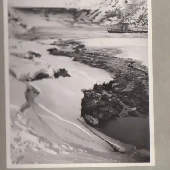 Ein Schwarz-Weiß-Foto zeigt eine verschneite Landschaft mit einer kleinen Hütte in der Nähe eines Gewässers. Im Hintergrund sind schneebedeckte Berge zu sehen.