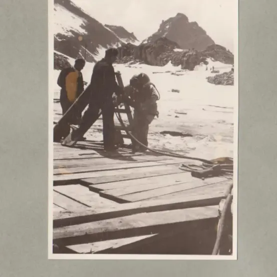 Drei Personen arbeiten auf einer hölzernen Plattform in einem verschneiten Gebirgsgebiet. Sie tragen Jacken und Stiefel. Eine Person hält eine Pfeife.