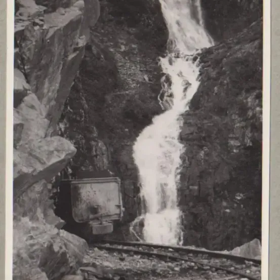 Ein Schwarz-Weiß-Bild zeigt einen Wasserfall, der einen steilen Felsvorsprung hinunterfließt, mit einem Bahngleis im Vordergrund.