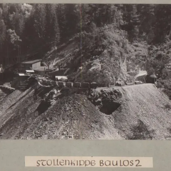 Ein Schwarz-Weiß-Foto eines Baugeländes mit einem kleinen Gebäude, Maschinen und Arbeitern. Der Bereich ist von Bäumen und felsigem Gelände umgeben. Der Text lautet 'STOLLENKIPPE BAU LOS 2'.