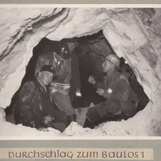 Drei Männer sitzen in einem Tunnel. Sie tragen Helme und Uniformen. Einer hält eine Lampe. Der Tunnel besteht aus Fels. Das Label lautet 'Durchschlag zum Baulos 1.'