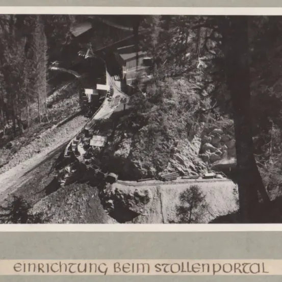 Ein Schwarz-Weiß-Bild einer im Bau befindlichen Straße in einem bewaldeten Gebiet, mit einem sichtbaren Tunnelportal. Bäume und Vegetation umgeben den Standort.