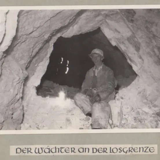 Ein Mann sitzt in einer Höhle, trägt einen Helm und hält eine Spitzhacke. Der Eingang der Höhle ist weit geöffnet und zeigt ein dunkles Inneres.