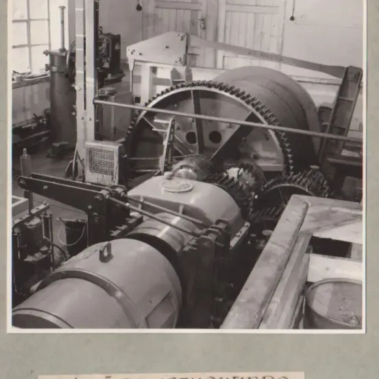 Ein Schwarz-Weiß-Foto einer industriellen Maschine mit großen Zahnrädern und einer Metallstruktur. Die Maschine befindet sich in einem Raum mit anderen Geräten.