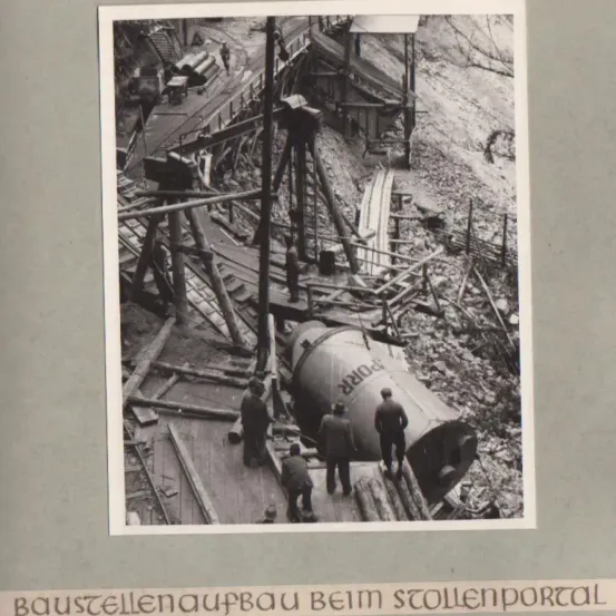 Ein Schwarz-Weiß-Foto zeigt Arbeiter auf einer Baustelle mit einer großen Röhre, die mit 'Krupp' beschriftet ist. Arbeiter stehen auf einer Plattform, einer kniet. Hölzerne Strukturen und Trümmer umgeben sie.