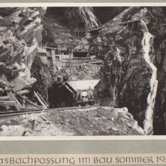 Schwarz-Weiß-Foto einer Baustelle in einem Gebirgsgebiet. Ein großer Kipplaster ist sichtbar, und eine Person steht in der Nähe.