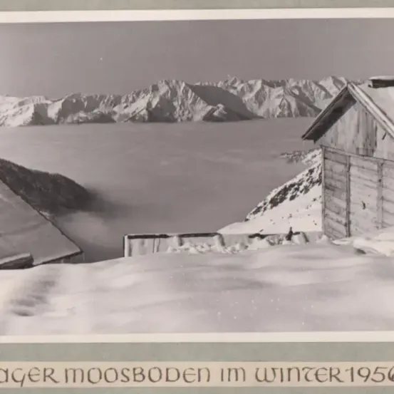 Ein Schwarz-Weiß-Foto einer Winterlandschaft mit einem Haus und einer Bergkette im Hintergrund. Schnee bedeckt den Boden und das Dach des Hauses. Das Bild hat einen Wasserzeichen am unteren Rand.