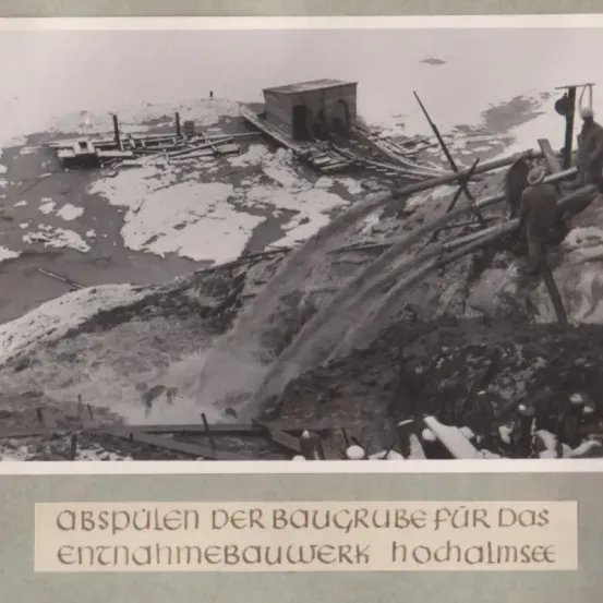 Ein Schwarz-Weiß-Foto zeigt eine Baustelle am Hochalmsee mit Arbeitern in Winterkleidung, einem provisorischen Gebäude und einem Wasserfluss.