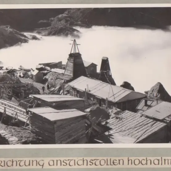 Ein Schwarz-Weiß-Foto zeigt eine bergige Landschaft mit verschiedenen Gebäuden und Strukturen, darunter ein großes Holzhaus und mehrere kleinere. Es gibt auch Gleise und Pfade, die durch das Gebiet führen.