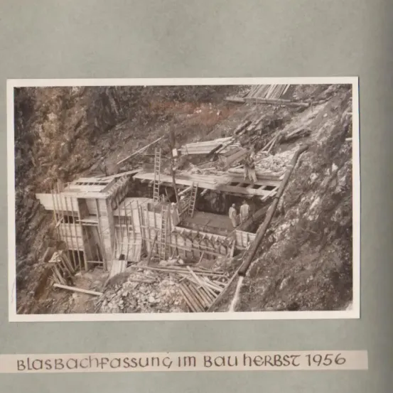 Schwarz-Weiß-Bild einer Baustelle im Bergtal. Arbeiter sind auf Gerüsten sichtbar, und der Text am unteren Rand lautet 'Blasbachpassage im Bau Herbst 1956'.