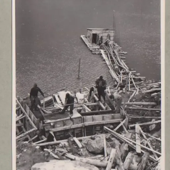 Ein Schwarz-Weiß-Foto von Menschen, die an einer Holzstruktur am Wasser arbeiten, mit Trümmern und Felsen drumherum.