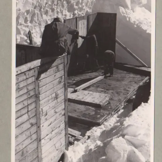 Ein Schwarz-Weiß-Bild zeigt Menschen, die an einer Holzstruktur in einer verschneiten Umgebung arbeiten. Drei Personen sind sichtbar, zwei beugen sich nach vorne, während die dritte in der Nähe einer Holzwand steht.