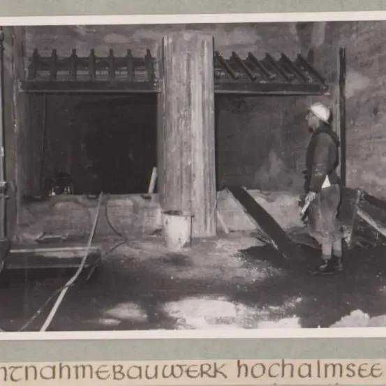 Ein altes Schwarz-Weiß-Bild zeigt einen Mann an einer Baustelle, der in der Nähe eines Eimers steht. Die Szene umfasst eine Steinmauer und eine Holzstruktur.