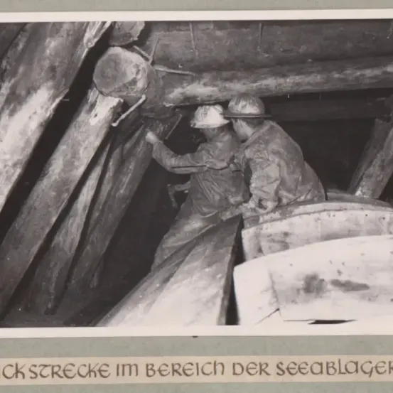 Schwarz-Weiß-Foto zeigt zwei Bergleute in einer Mine, beide tragen Helme und arbeiten in einem Tunnel. Der Tunnel wird von Holzbalken gestützt.