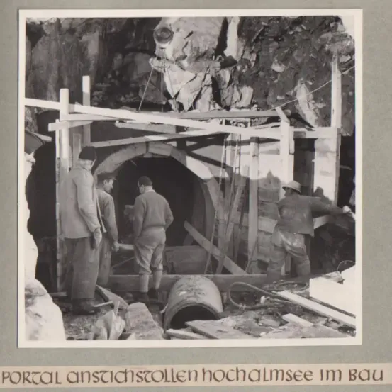 Ein Schwarz-Weiß-Foto von Arbeitern in einem Tunnel mit sichtbaren Trümmern und einer großen Rohrleitung im Vordergrund.