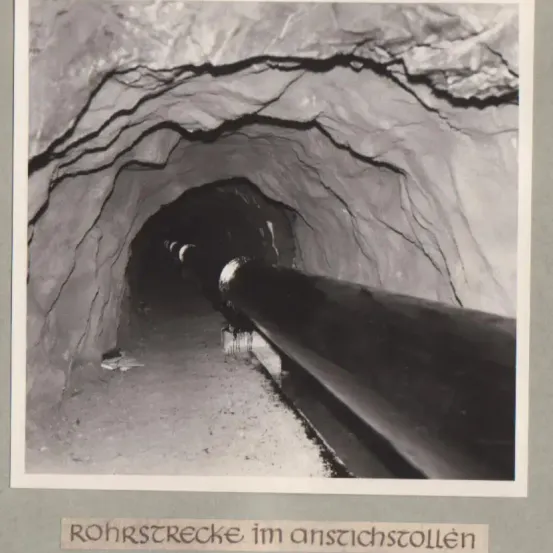 Ein Schwarz-Weiß-Bild zeigt einen langen Tunnel mit einem großen Rohr in der Mitte. Die Tunnelwände sind ungleichmäßig, und das Rohr scheint in der Erde vergraben zu sein.