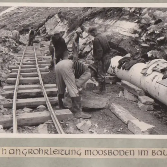 Schwarz-Weiß-Foto zeigt Arbeiter bei der Verlegung von Bahngleisen. Ein Mann kniet, ein anderer steht und mehrere gehen. Im Hintergrund befinden sich große Steine und eine Rohrleitung.