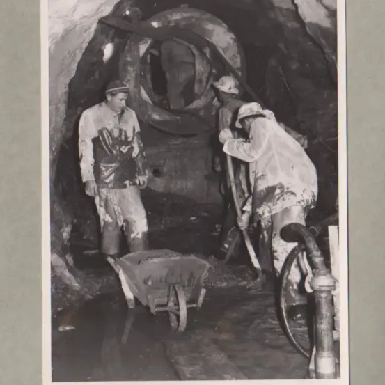 Schwarz-Weiß-Bild von drei Bergleuten in einem Tunnel. Einer schiebt einen Schubkarren, während die anderen Werkzeuge halten.