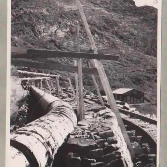 Ein Schwarz-Weiß-Bild zeigt einen Berg mit einer großen Holzkonstruktion und einer großen Rohrleitung am Boden.