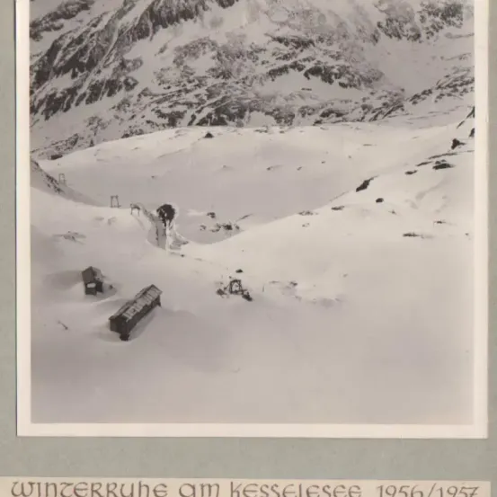 Ein Schwarz-Weiß-Foto einer verschneiten Berglandschaft mit zwei kleinen Gebäuden. Das Bild trägt den Titel 'Winterruhe am Kesselsee 1956/1957'.