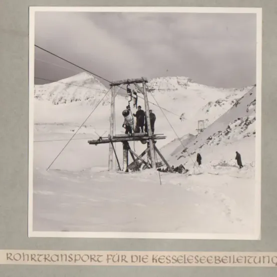 Ein Schwarz-Weiß-Foto zeigt eine Seilbahnstruktur in einer verschneiten Bergumgebung. Einige Personen stehen auf der Plattform der Seilbahn.