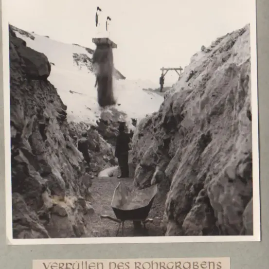 Ein Schwarz-Weiß-Foto zeigt einen Mann, der in einem Tunnel steht. Er trägt einen Hut und hält eine Schubkarre. Schneebedeckte Felswände umgeben ihn. Der Tunnel hat eine Holzkonstruktion und eine Rohrleitung. Das Foto trägt den Titel 'Verfüllen des Rohrgrabens.'