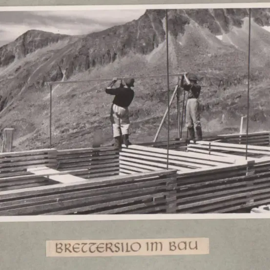Schwarz-Weiß-Foto von zwei Männern, die auf einer Baustelle in einem Berggebiet arbeiten. Ein Mann hält ein Vermessungsinstrument.