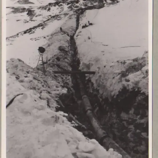 Ein Schwarz-Weiß-Foto zeigt eine tiefe Schlucht im verschneiten Gelände mit einem großen Rohr, das hindurchläuft. Ein Stativ mit einer Kamera ist in der Nähe positioniert.