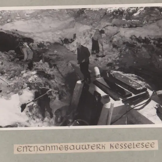 Ein Schwarz-Weiß-Bild zeigt Arbeiter in Winterkleidung, die in einem verschneiten Gebirgsgebiet graben und arbeiten.
