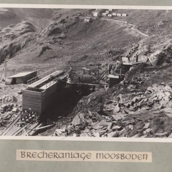 Ein Schwarz-Weiß-Foto von Brecheranlage Moosboden, einer Baustelle mit Maschinen, Menschen und verstreuten Materialien an einem Berghang.