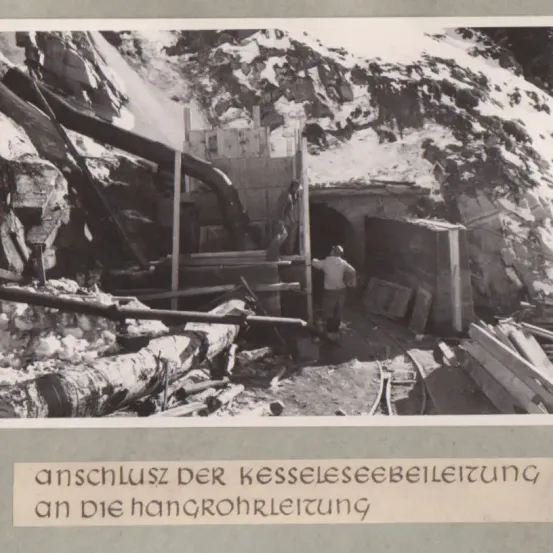 Ein Schwarz-Weiß-Foto zeigt eine Baustelle mit einem Tunneleingang, einer Person im Vordergrund und verschiedenen Holzkonstruktionen. Im Hintergrund sind schneebedeckte Berge zu sehen.