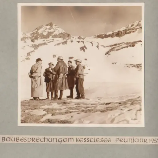 Eine Gruppe von Männern in Mänteln und Mützen steht in einem verschneiten Gebirgsgebiet. Sie diskutieren etwas und schauen in die gleiche Richtung. Das Bild ist mit dem Text 'Baubesprechung am Kesselsee - Fruhjahr 1957' beschriftet.