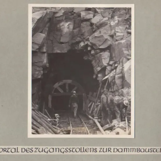Ein Schwarz-Weiß-Foto zeigt den Eingang eines Tunnels mit Arbeitern. Ein Mann geht durch den Tunnel, andere arbeiten am Tunneleingang.