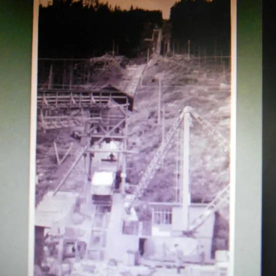 Ein monochromes Bild zeigt eine Baustelle mit einer Holzstruktur und Maschinen. Bäume und ein geneigtes Gelände sind im Hintergrund zu sehen.