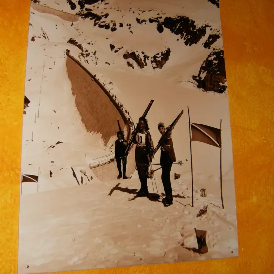 Ein sepiafarbenes Foto zeigt drei Skifahrer, die im Schnee stehen. Sie halten Skistöcke und tragen Winterkleidung. Rechts von ihnen steht eine Fahne.