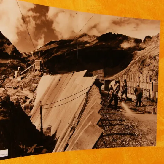 Ein altes Foto zeigt Arbeiter in Schutzhelmen, die auf einem bergigen Gelände mit Bauarbeiten beschäftigt sind, umgeben von Drahtzäunen und mit Maschinen im Hintergrund.